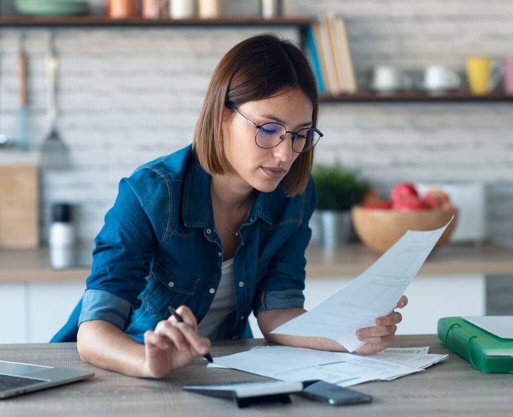 Mujer joven con gafas revisando documentos financieros en una cocina moderna, concentrada mientras toma notas con una pluma.