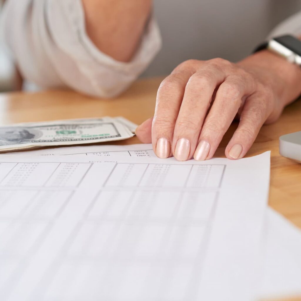 Mano de una persona tocando una hoja de cálculo con tablas financieras, con billetes de dólar y una laptop parcialmente visibles sobre la mesa.