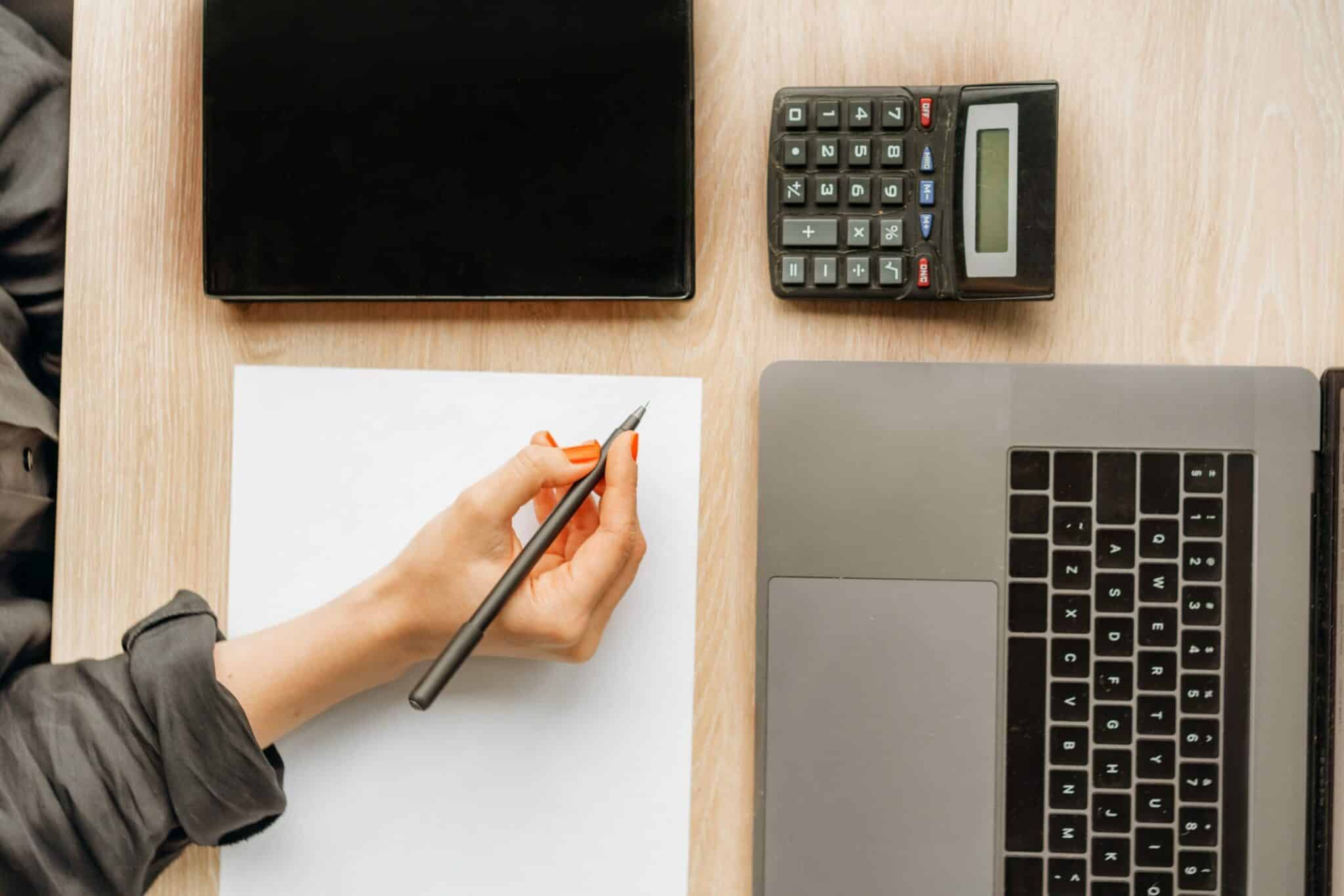 Mesa vista de cima com laptop, calculadora, pasta e folha de papel com uma mão sobre segurando uma caneta.