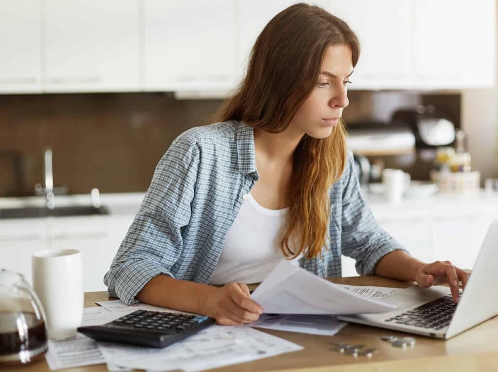 Mulher analisando documentos e usando um laptop na cozinha, concentrada em tarefas financeiras ou contábeis.