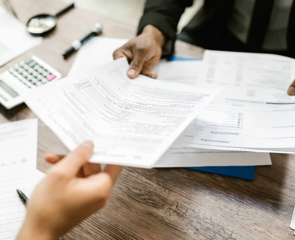 Duas pessoas revisando e trocando documentos fiscais ou contábeis sobre uma mesa, com calculadora e papéis ao fundo.