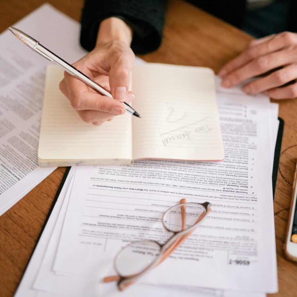 Pessoa escrevendo em caderno sobre mesa de madeira com formulários na sua volta e calculadora aberta no celular