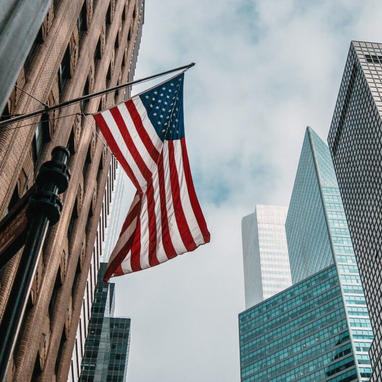 USA flagpole near skyscrapers with cloudly sky.