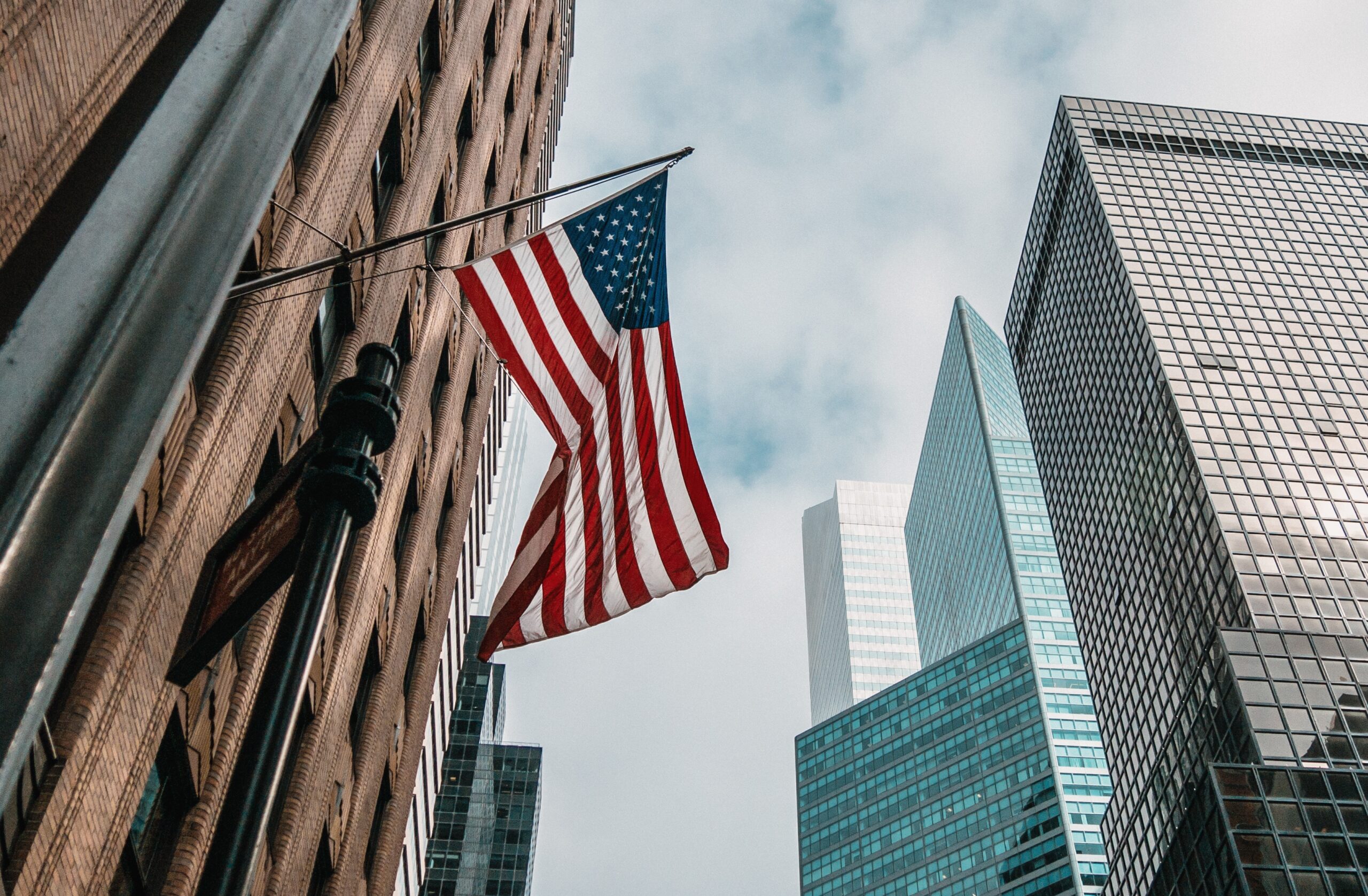 USA flagpole near skyscrapers with cloudly sky.