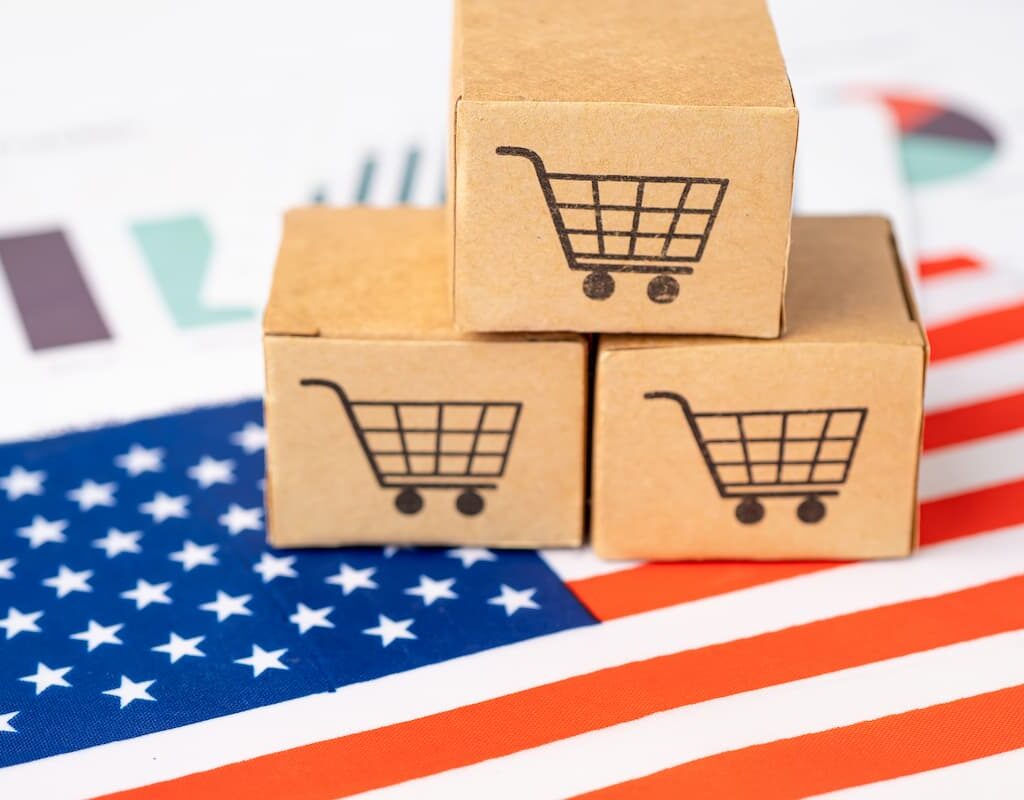 Three cardboard boxes with shopping cart icons stacked together on top of a U.S. flag and business charts, symbolizing e-commerce in the United States.