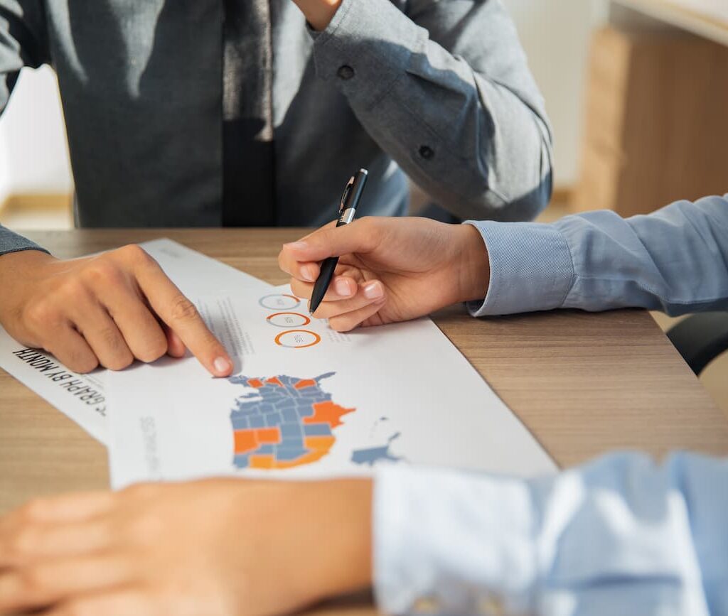 Close-up of two people reviewing a U.S. map with color-coded states on a printed document, discussing data and pointing with a pen.