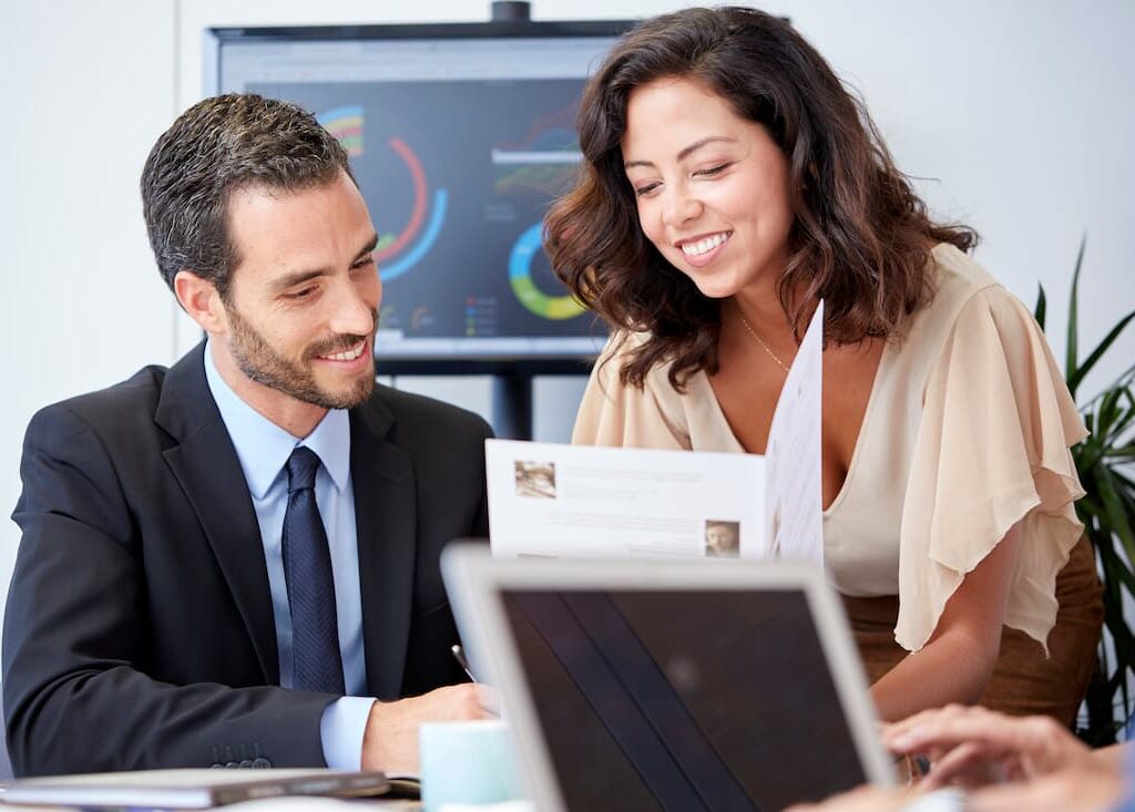 Two professionals smiling and reviewing a printed document together in an office setting, with data charts displayed on a screen in the background.