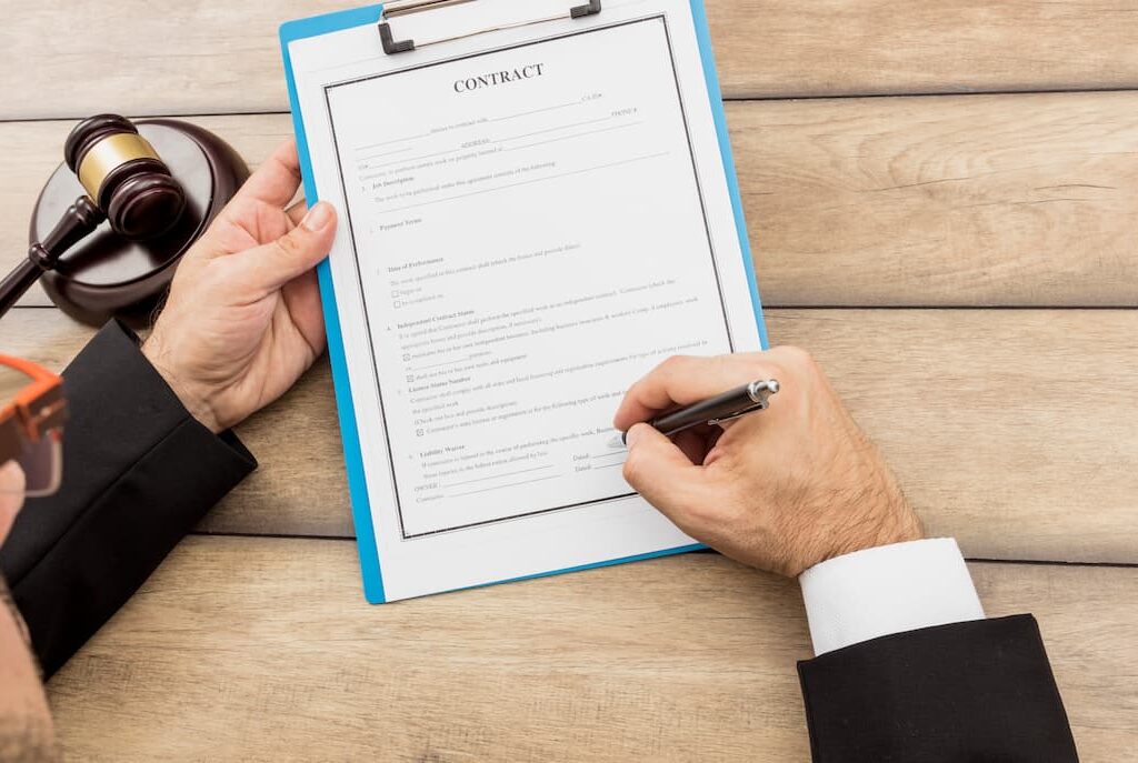Person in formal attire signing a printed contract on a clipboard, with a judge's gavel placed on the wooden desk beside them.