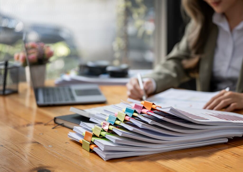 Stacks of organized documents clipped with colorful binders on a wooden desk, with a businesswoman working in the background near a laptop and window.