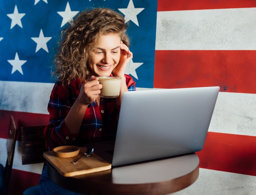 Smiling woman holding a cup of coffee while using a laptop, sitting in front of a large U.S. flag backdrop.