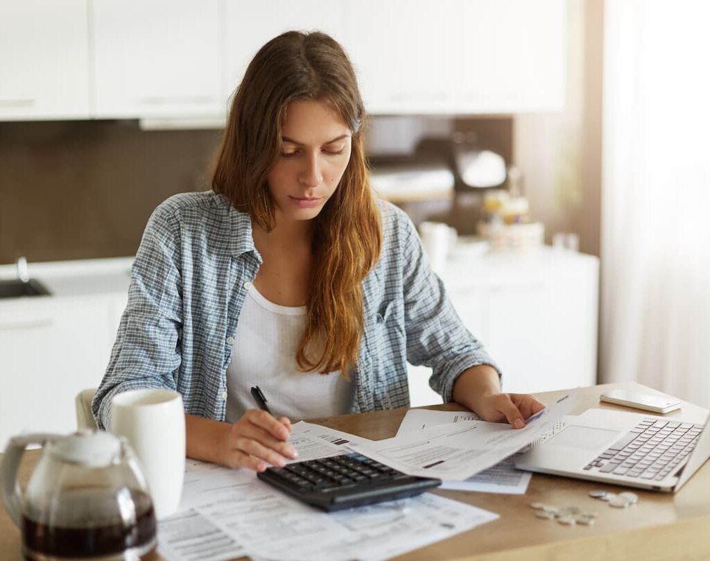 Mujer trabajando en una mesa con documentos y una calculadora, revisando papeles mientras utiliza su laptop en un ambiente doméstico.