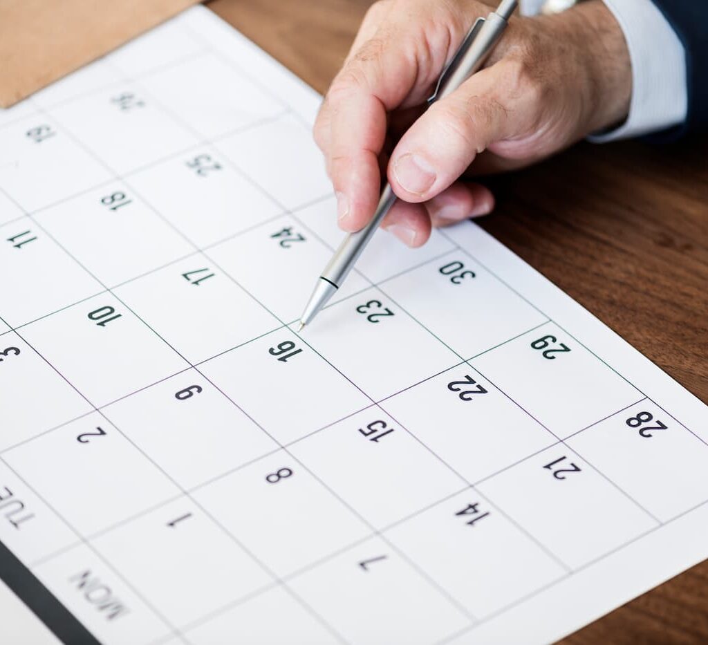 Primer plano de una mano que sostiene un bolígrafo y señala una fecha específica en un calendario colocado sobre un escritorio de madera.