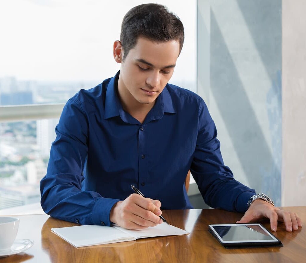Un joven con una camisa azul escribe en un cuaderno en un escritorio, con una tableta y una taza de café cerca, en una oficina de gran altura.