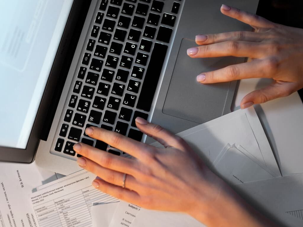 Persona escribiendo en una computadora portátil con documentos dispersos por todas partes, sugiriendo ingresar datos o trabajar en papeleo.