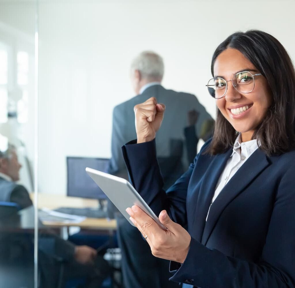 Una mujer de negocios sonriente con traje sostiene una tableta y levanta el puño en señal de celebración, mientras sus colegas trabajan en el fondo de una oficina.