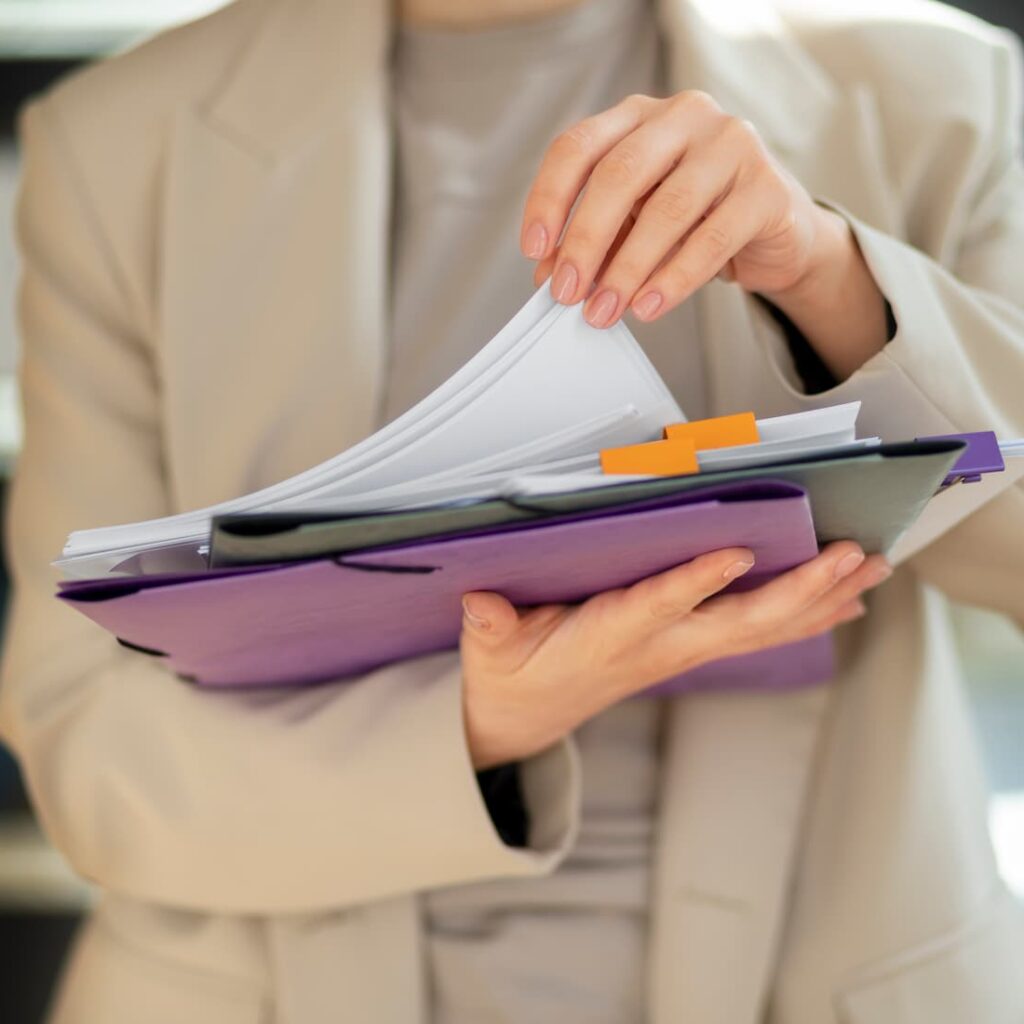 Person in a beige blazer flipping through documents in a folder, with several pages and colored tabs visible.