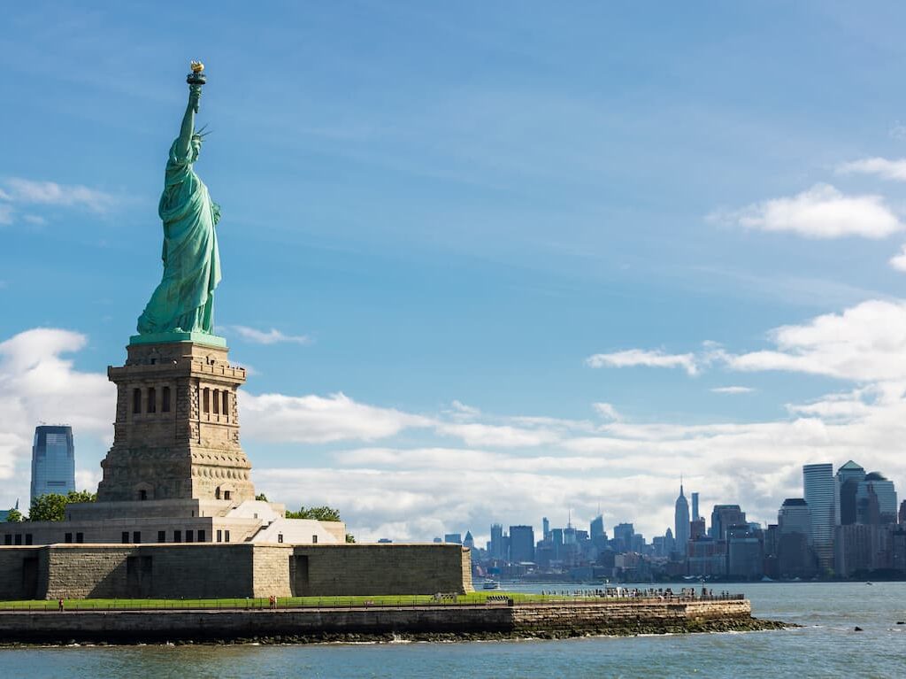 The Statue of Liberty standing tall on Liberty Island with the New York City skyline visible in the background on a clear day.
