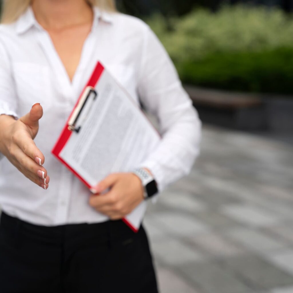 Woman extending her hand for a handshake while holding a clipboard with documents, suggesting a business agreement or professional introduction outdoors.