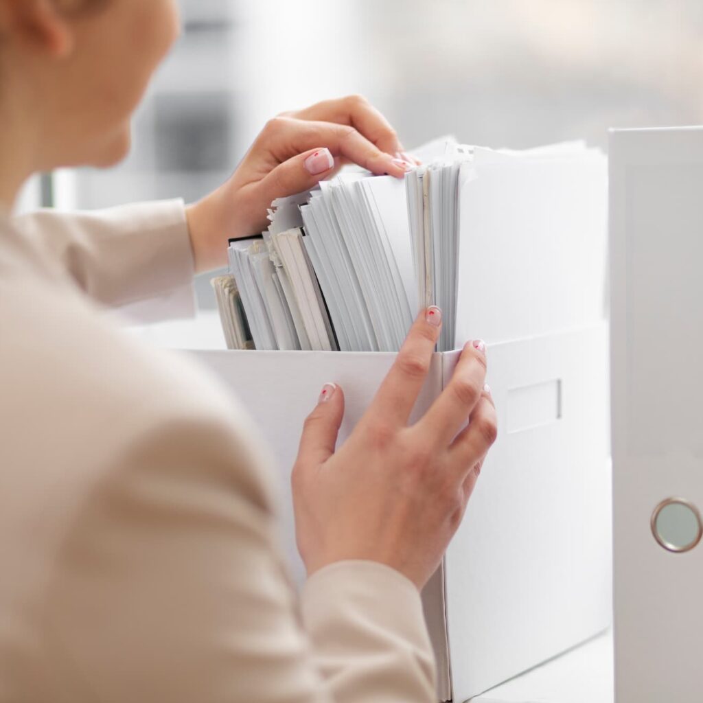 Person organizing or searching through a white file box filled with documents, suggesting paperwork management or office filing tasks.