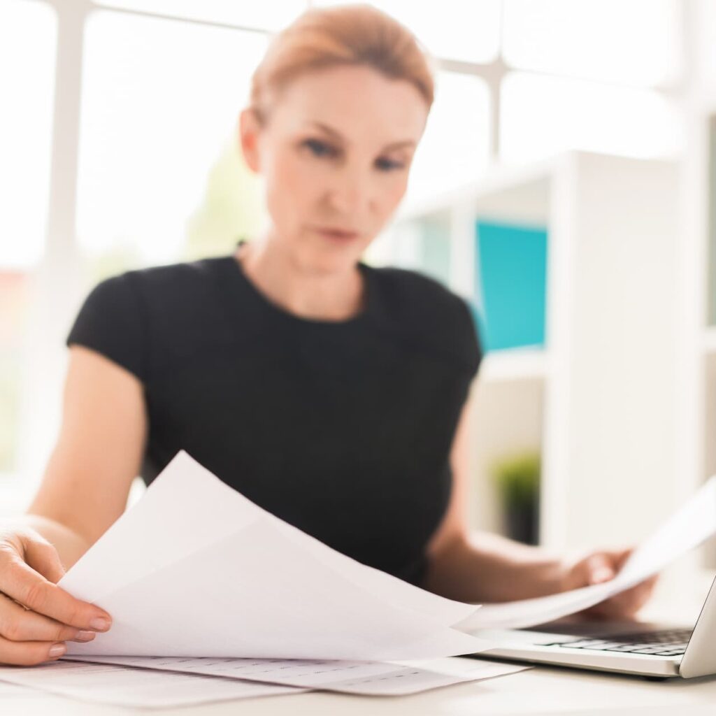 Woman in a black top reviewing documents at her desk in a bright office, with focus on the papers in the foreground.